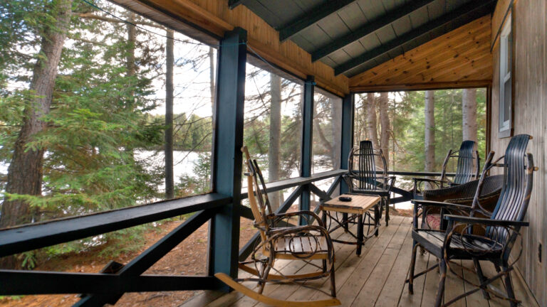four rustic wooden chairs and a coffee table on a rustic screened porch of a cabin sitting in a coniferous forest with a view of water