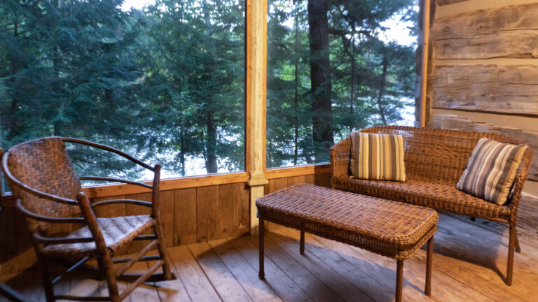 a wicker chair, love seat and table inside a screened in covered porch of a log cabin overlooking a lake and coniferous trees