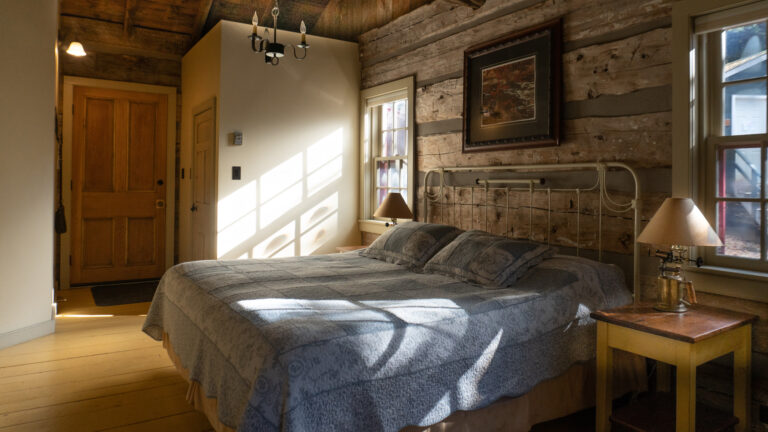 a sun-filled bedroom in a log cabin with a large bed, blue comforter, wooder side table and antique brass lamp