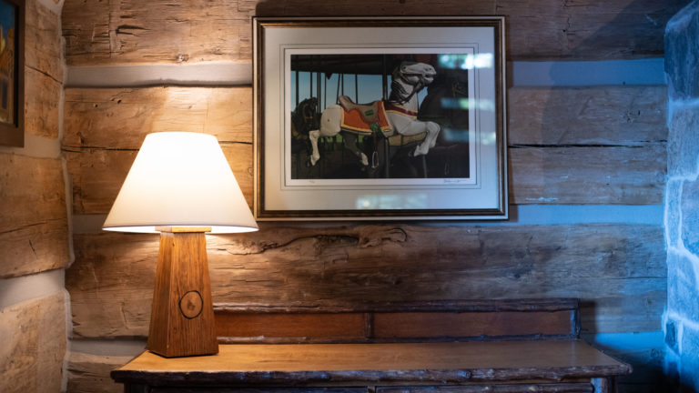vignette of a rustic wooden illuminated lamp on a live edge wooden surface with a merry go round photograph hanging on the wall of a log cabin