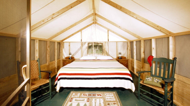 inside of a wood and canvas tent with two green wooden chairs, two antique wooden sidetables with lanterns and a bed covered in a Hudson's Bay point blanket