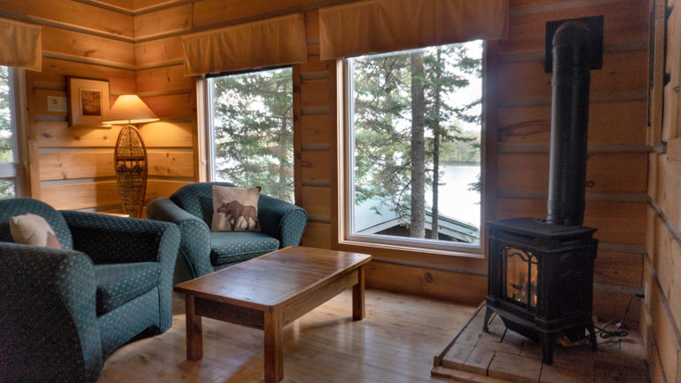 two green upholstered chairs with moose pillows in a wood-paneled living room with a wood burning stove and wooden coffee table with a view of a lake and pine trees through large windows