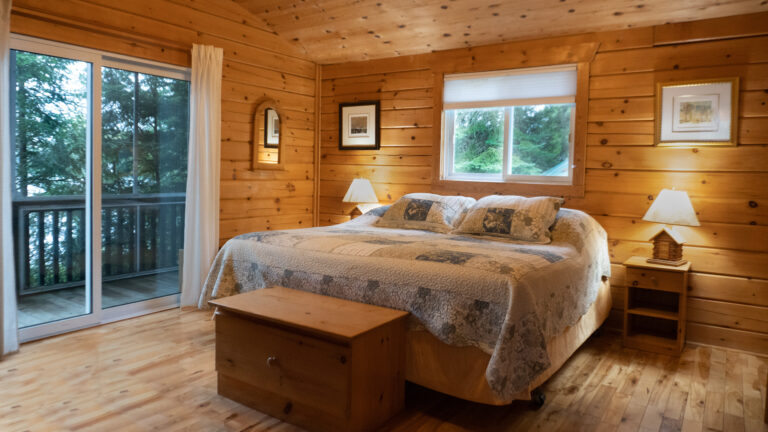 large bed in the middle of a wood paneled room flanked by side tables with lamps and a wooden chest at the foot of the bed. View of coniferous forest and water is visible through glass sliding doors