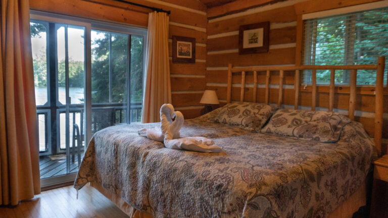 large bed with a neutral floral quilt, log headboard and two towels folded like swans in a loving embrace in a log cabin with a view of the lake through glass paneled sliding doors