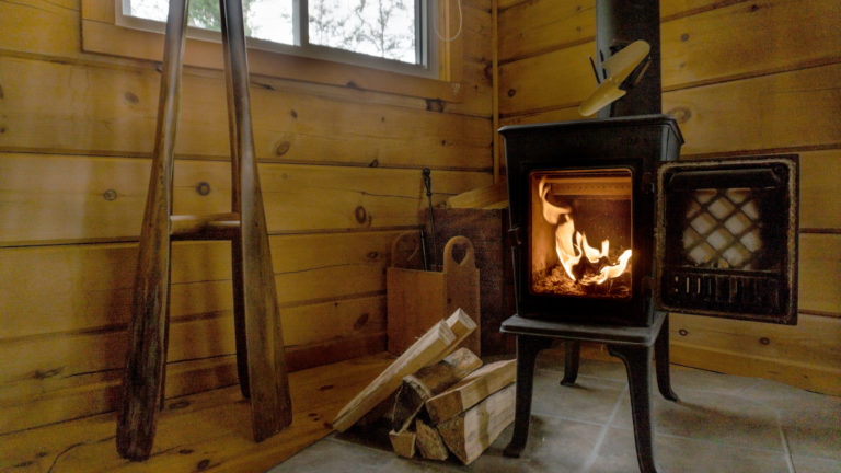 lit standalone cast iron fireplace with door open inside a log cabin with a pile of chopped wood sitting beside it