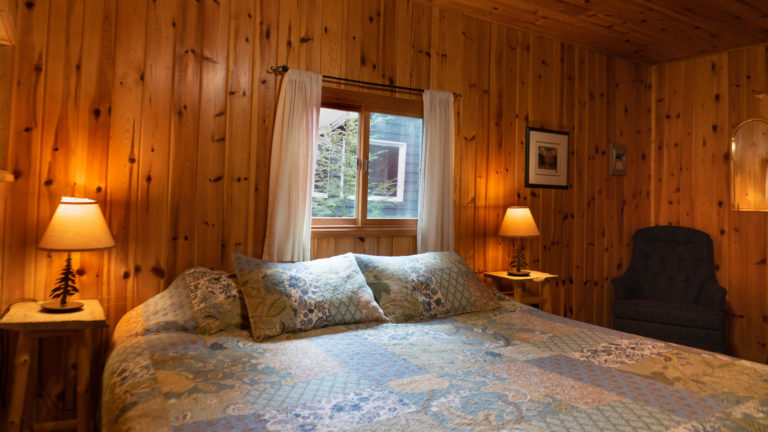 large bed in a wood paneled room with blue patch work quilt, blue easy chair and two side tables with lit lamps