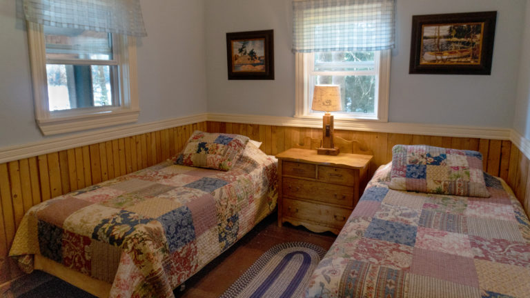 two twin beds covered in patch work quilts seperated by an antique dresser with lamp insude a blue and wood paneled room
