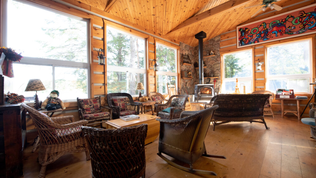 sun-filled room with pine paneling on the walls filled with wicker lounge furniture, a wood burning stove and indigenous Canadian artwork